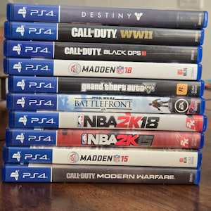 Può includere: Una pila di custodie di videogiochi per PS4, tra cui titoli come "Destiny", "Call of Duty: WWII" e "Madden 18". Le custodie sono principalmente nere, bianche e blu, con i titoli dei giochi stampati sui dorsi.