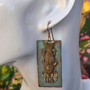May include: A rectangular earring with a copper-coloured frame and a turquoise background. A brass fish is centered on the earring, with a detailed design. The earring is suspended from a copper-coloured hook.