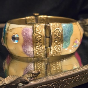 May include: A hinged bangle bracelet with a cream-colored enamel base, accented with gold-tone metal and colorful sections of blue, purple, and yellow. The bracelet features a decorative clasp and is embellished with clear, round rhinestones.