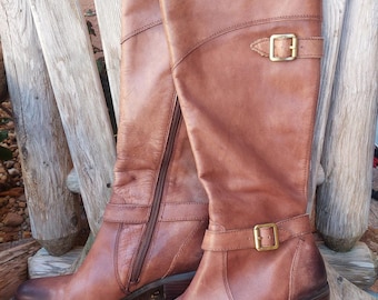 May include: A pair of knee-high brown leather boots with a side zipper and buckle detail. The boots have a slight heel and are displayed on a wooden surface. The leather has a distressed appearance.