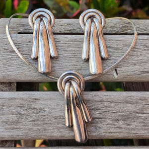 May include: A silver-toned jewelry set featuring a necklace and matching earrings. The necklace is a thin, flexible band with a pendant. The earrings and pendant have a knotted design with three elongated, rounded elements. The set is displayed on a wooden surface.