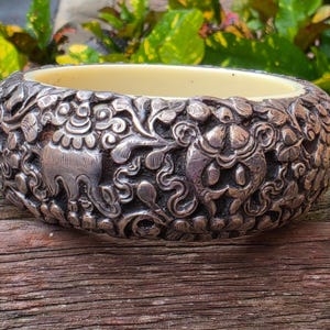 May include: A silver-toned bangle bracelet with intricate floral and animal carvings. The interior is a light cream color. The bracelet is resting on a weathered wooden surface, with green foliage in the background.