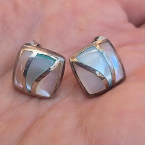 May include: Two square silver earrings with iridescent mother-of-pearl inlays. The earrings have a geometric design with silver accents and a post backing. The inlays feature shades of pink, blue, and white, creating a shimmering effect.