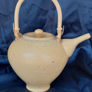 May include: A light yellow ceramic teapot with a bamboo handle and lid. The teapot has a rounded body and a spout on the side.