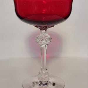 May include: A red glass goblet with a wide bowl and a clear, ornate stem. The bowl is a deep red colour, and the stem features decorative detailing. The goblet sits on a clear, circular base. The glass is transparent, allowing light to pass through.