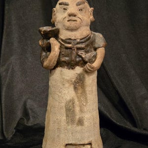 May include: A weathered, antique ceramic figurine of a man holding a bowl and a small object. The figure is dressed in a tunic and has a detailed face with a mustache and beard. The color palette is primarily earthy tones.