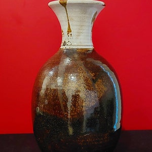 May include: A handcrafted ceramic vase with a bulbous body and a flared neck. The vase features a speckled brown glaze that transitions to a dark brown base. The neck is white with brown drips.