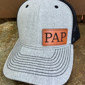 May include: A gray and black trucker hat with a brown leather patch that says "PAP" on it.
