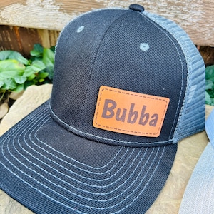 May include: A black and gray trucker hat with a brown leather patch that says "Bubba" on it.