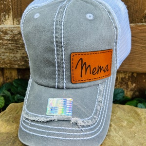 May include: Gray and white distressed trucker hat with a brown leather patch that says "Mema" on it.