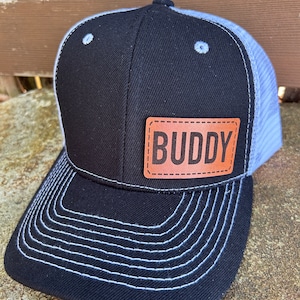 May include: Black and blue trucker hat with a brown leather patch that says "BUDDY" in white stitching.