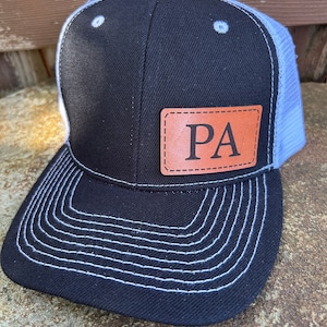 May include: A black and blue trucker hat with a brown leather patch that says "PA" on it. The hat has a white stitching detail around the brim.