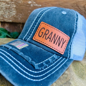 May include: A blue denim trucker hat with a light blue mesh back. The hat has a distressed look with frayed edges and white stitching. A brown leather patch with the word "Granny" is sewn onto the front of the hat.
