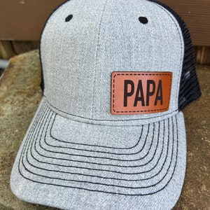 May include: A gray and black trucker hat with a brown leather patch that says "PAPA" on it.