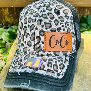 May include: A black and brown leopard print trucker hat with a brown leather patch that says "CoCo" on it. The hat has a distressed look and a mesh back.