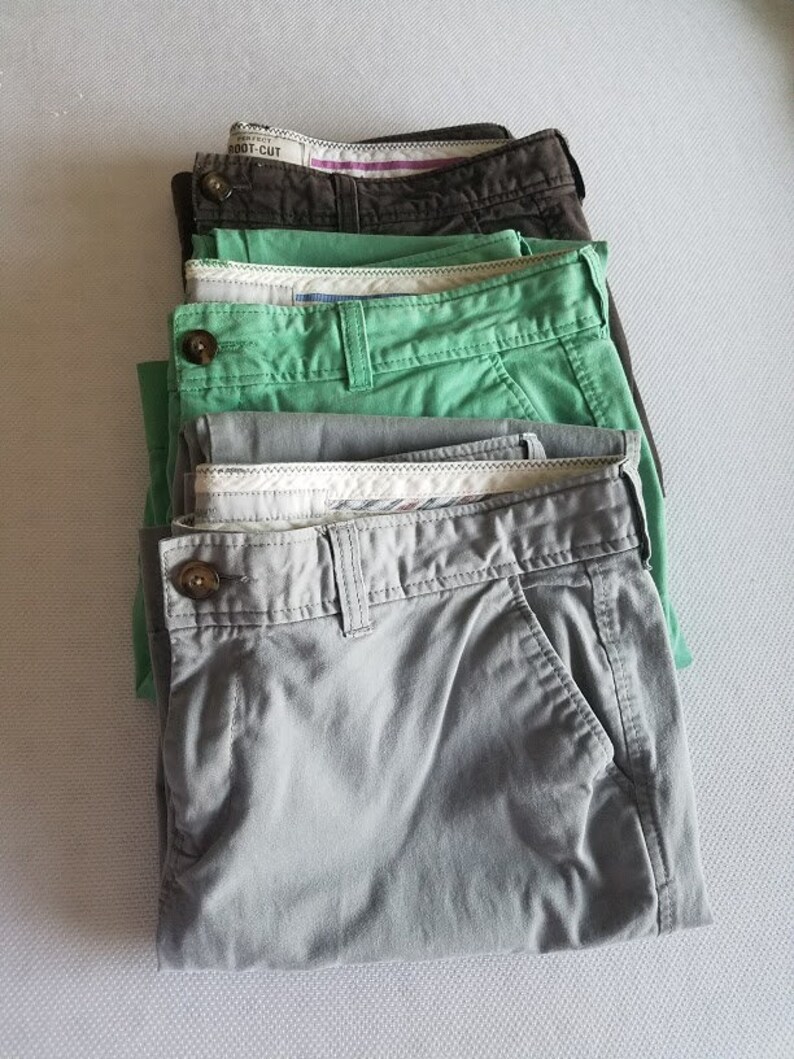 May include: A stack of five pairs of khaki pants in different shades of green and gray. The pants are folded and stacked on top of each other. The top pair of pants has the text "BOOT-CUT" printed on the waistband.