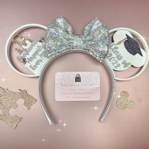 Peut inclure: Des oreilles de Minnie Mouse blanches avec un nœud en paillettes argentées. L'oreille gauche a un château blanc avec le texte "Félicitations" et "Chaque moment magique". L'oreille droite a un bonnet de graduation blanc avec le texte "Classe de 2022".