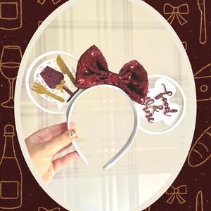 May include: White headband with mouse ears, featuring a sequined burgundy bow. One ear has a fork, knife, and wine glass design, the other ear has the text "Food & Wine". The background is a burgundy colour with gold illustrations.
