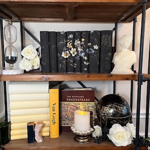 Puede incluir: Una estantería de madera con dos estantes, decorada con libros, un globo terráqueo, una vela y flores blancas. Los libros están apilados en el estante inferior, con un globo terráqueo y una vela delante de ellos. El estante superior tiene una pila de libros con un diseño floral en las cubiertas. Hay flores blancas en ambos estantes.