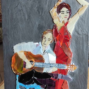 Op de afbeelding: Mozaïek kunstwerk met een flamencodanseres in een rode jurk en een gitarist. Het kunstwerk is gemaakt op een donkergrijze leisteen, met de figuren gemaakt van kleurrijke glasstukken. De gitarist zit en speelt een bruine gitaar.