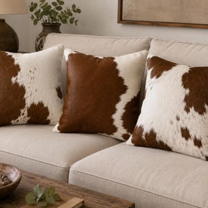 May include: Three square throw pillows featuring a brown and white cowhide pattern. Each pillow has a textured, hair-on-hide appearance with irregular patches of dark brown and cream white fur distributed across the front surface of the cushions.