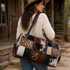 Op de afbeelding: Een patchwork leren duffeltas met een mix van zwarte, witte, bruine en bruingele koeienhuid vierkanten. De tas heeft bruine leren handvatten en een schouderriem. De tas wordt over de schouder gedragen.