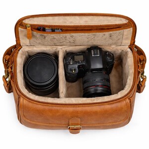 Può includere: Borsa per fotocamera in pelle marrone chiaro aperta, con cerniera superiore e fibbia color oro. L'interno ha due scomparti, uno con un obiettivo della fotocamera e l'altro con una fotocamera. La borsa ha una tracolla e una piccola tasca con cerniera.