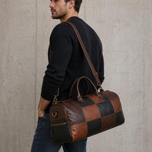 Op de afbeelding: Een bruine, zwarte en tan leren duffeltas met een patchwork ontwerp. De tas heeft een schouderriem en handvatten. De tas heeft een ritsvak en goudkleurige hardware.