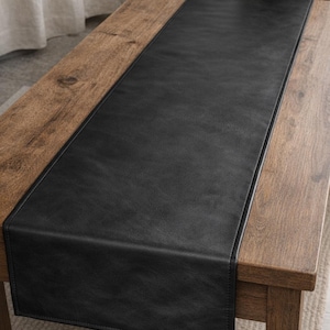 Puede incluir: Un camino de mesa de cuero negro está centrado en una mesa de madera. El camino de mesa es rectangular y tiene un borde cosido. La mesa es de madera de color marrón claro. Un jarrón con flores blancas y bolas decorativas está sobre la mesa.