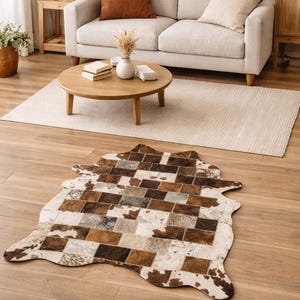 May include: A cowhide rug with a patchwork design in shades of brown, tan, and white. The rug is irregularly shaped and lies on a hardwood floor. The room also features a beige sofa, a round wooden coffee table, and a neutral-colored rug.