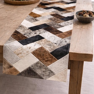May include: A rectangular table runner with a geometric pattern of brown, black, and cream-coloured cowhide patches. The runner is placed on a wooden table. Candles and a decorative bowl are also on the table.