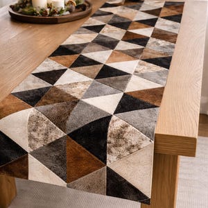 May include: A decorative table runner with a geometric patchwork design. The runner features triangles in shades of brown, black, grey, and cream. The runner is placed on a wooden table, with candles and a decorative tray in the background.