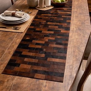 May include: A rectangular table runner with a patchwork design of brown and black leather rectangles. The runner is placed on a wooden table, with place settings and decorative items visible.