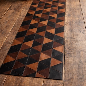 May include: A geometric table runner with a repeating pattern of black, brown, and tan triangles. The runner is made of leather or a leather-like material and is placed on a wooden table. The runner is approximately 60 inches long.
