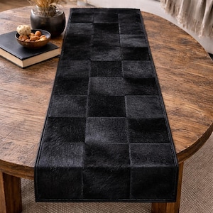 May include: A black rectangular table runner with a checkerboard pattern made of textured material. The runner is placed on a round wooden table, adding a touch of elegance to the home decor.