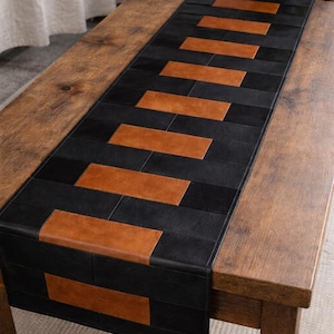 May include: A rectangular table runner with a geometric pattern of black and brown leather rectangles. The runner is placed on a wooden table. The color scheme is neutral, with a focus on texture and design.