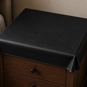 May include: A black, rectangular table cover draped over a wooden nightstand. The cover has a textured surface and hangs over the edges of the nightstand. The nightstand has two drawers with black knobs.