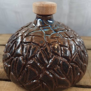 Op de afbeelding: Een bruine keramische fles met een getextureerd oppervlak en een houten deksel. De fles heeft een unieke, bijna gebarsten glazuur.