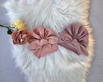 May include: Three dusty rose colored turban headbands with a knotted bow on top. The headbands are on a white fluffy surface.