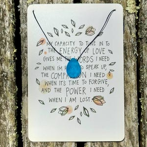 May include: A teardrop-shaped, light blue gemstone pendant on a black cord necklace. The pendant is displayed on a white card with handwritten text and floral illustrations. The card's message is about love and inner strength.