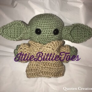 May include: A crocheted green and tan plush toy resembling a character from the Star Wars franchise. The toy has large eyes and a small mouth. The text "Ittie Bittie Toes" is visible on the toy's chest.