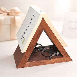 May include: A triangular wooden book stand with a book titled "Blink" and a pair of eyeglasses. The stand is made of dark brown wood and has a natural finish. The glasses are black-framed.