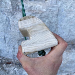 Op de afbeelding: Een houten klimgreep met een touw eraan bevestigd. De greep is lichtbruin en heeft een unieke vorm.