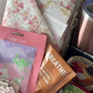 Close-up of blind date with a book box showing wrapped floral book, annotation tabs, tumbler, tea, and self care items. Mother’s Day gift for mom, cozy reading care package, and book lover gift set. Mystery book bundle designed for relaxing reading experiences, aesthetic bookish gifts, and thoughtful personalized book box ideas.