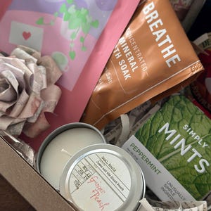 Close-up of Mother’s Day book box with candle, bath soak, tea, mints, and wrapped book arranged inside a curated blind date with a book gift set. Cozy self care box for readers featuring bookish accessories and relaxing essentials. Perfect gift for mom, book lover gift, mystery book bundle, and aesthetic reading care package for quiet nights and thoughtful gifting.