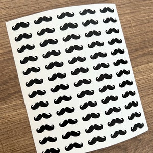 Puede incluir: Hoja de pegatinas blancas con un patrón repetido de bigotes negros. Los bigotes tienen un diseño clásico y curvado, espaciados uniformemente en la hoja de pegatinas. El fondo es una superficie de madera.