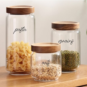 May include: Three glass jars with wooden lids, filled with various dry goods. The jars are labeled "pasta", "grains", and "oats".