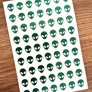 May include: A sheet of white paper with 50 green glitter alien head stickers. The stickers are arranged in a grid pattern.