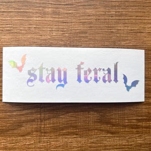 May include: A white sticker featuring the phrase "stay feral" in a gothic font with a holographic, iridescent finish. Two bat silhouettes are on either side of the text.