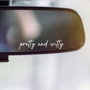 以下が含まれることがあります： 「pretty and witty」という文字が筆記体で書かれた車のバックミラー。ミラーは長方形で角が丸く、フレームは暗い色。背景はぼやけており、屋外のシーンを示唆している。